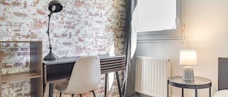 Una habitación con pared de ladrillo, un escritorio con silla, una lámpara y una mesita de noche con lámpara.