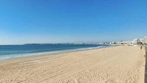 Una amplia playa de arena con cielo azul despejado, un mar tranquilo y edificios a lo lejos.