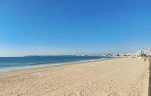 Ein weiter Sandstrand, strahlend blauer Himmel, ruhiges Meer und Gebäude in der Ferne.