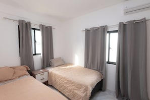 Ein Schlafzimmer mit zwei Betten, einem Nachttisch, einem Fenster mit Vorhängen und einer Klimaanlage.