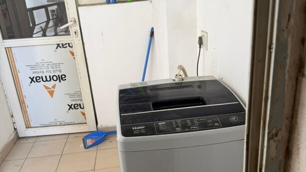 A room with a Haier washing machine, a broom, and a window with a view of a building. - Beautiful 3-bedroom apartment with AC in cool Noida neighborhood (Noida)