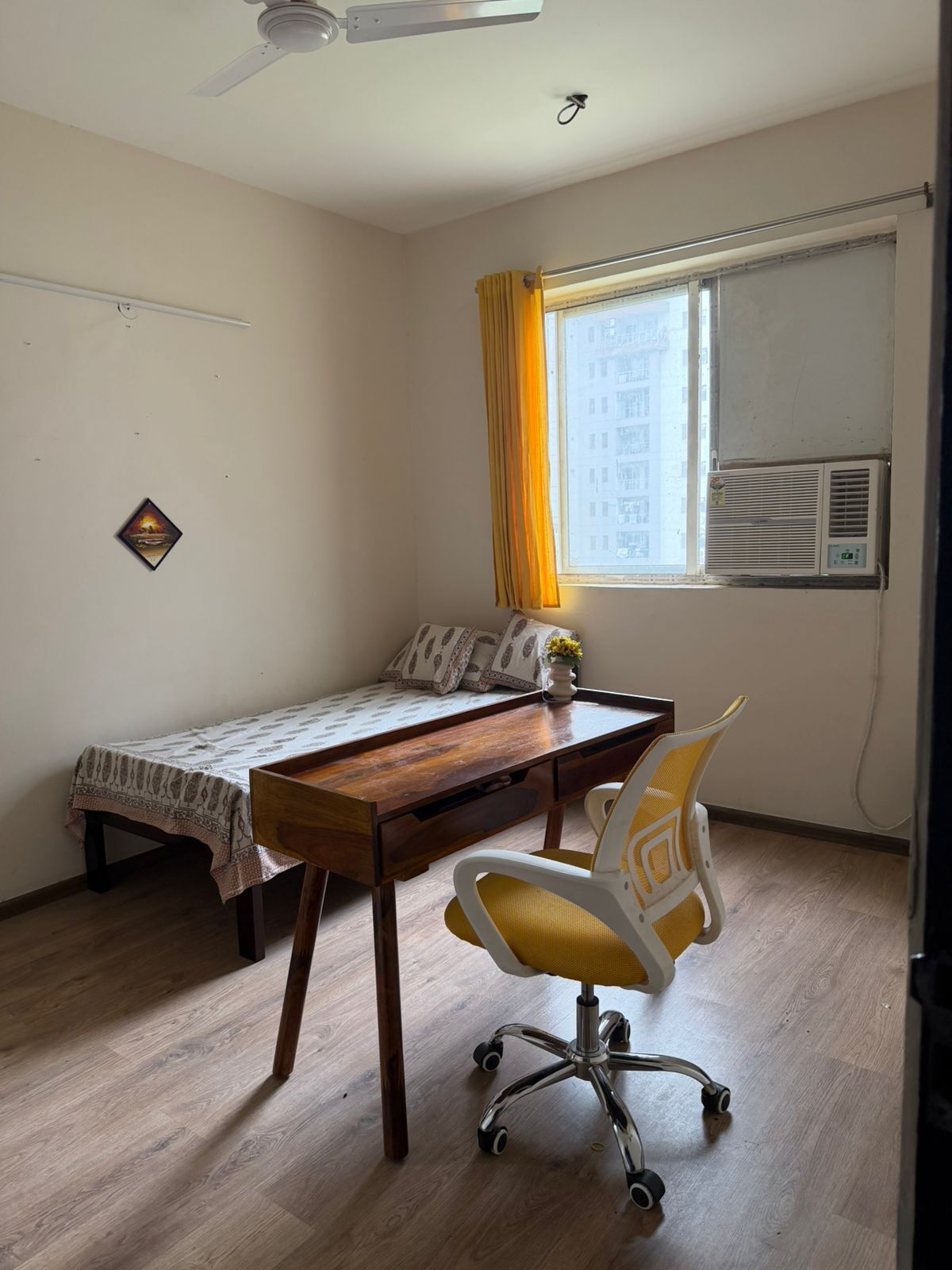Ein kleines Zimmer mit einem Bett, einem Schreibtisch, einem Stuhl, einem Deckenventilator, einem Fenster mit Vorhängen und einer Klimaanlage.