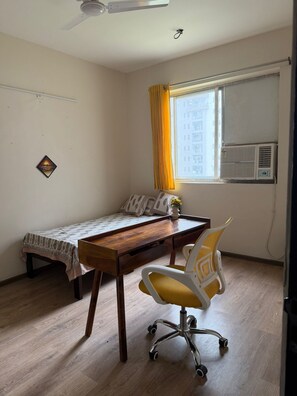 Ein kleines Zimmer mit einem Bett, einem Schreibtisch, einem Stuhl, einem Deckenventilator, einem Fenster mit Vorhängen und einer Klimaanlage.