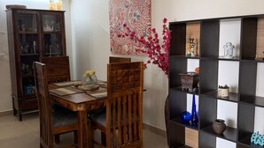 A dining area with a wooden table and chairs, a cabinet with glass doors, a ceiling fan, and a wall-mounted shelf with decorative items. - Beautiful 3-bedroom apartment with AC in cool Noida neighborhood (Noida)
