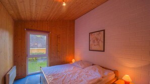 A bedroom with a wooden ceiling, a bed with a patterned bedspread, a nightstand with a lamp, and a window with a view of greenery. - Komfortabler Doppelbungalow mit Terrasse im Ferienpark Mirow (Mirow)