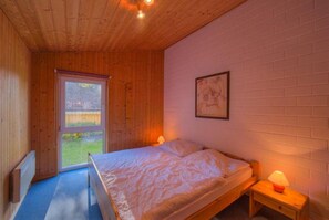 Ein Schlafzimmer mit Holzbalkendecke, einem Bett mit gemustertem Bettbezug, einem Nachttisch mit Lampe und einem Fenster mit Blick ins Grüne.