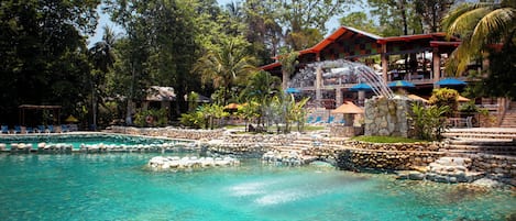 A pool with clear water, surrounded by rocks and a stone wall. There are lounge chairs and umbrellas in the background.