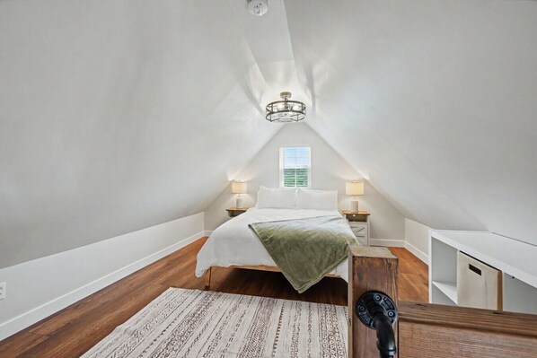 A bedroom with a sloped ceiling, a bed with a green blanket, two bedside lamps, and a window with blinds. - The Palmer - A Modern Cottage in Wine Country (Hermann)