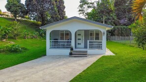 Ein kleines, eingeschossiges Haus mit Veranda und Eingangstür, umgeben von einem gepflegten Rasen und Bäumen.