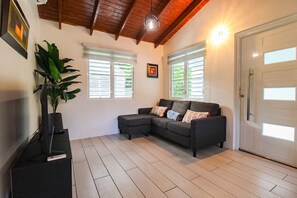 Ein Wohnzimmer mit einem dunkelgrauen Ecksofa, zwei Fenstern mit Jalousien, einer Zimmerpflanze und einer Tür.