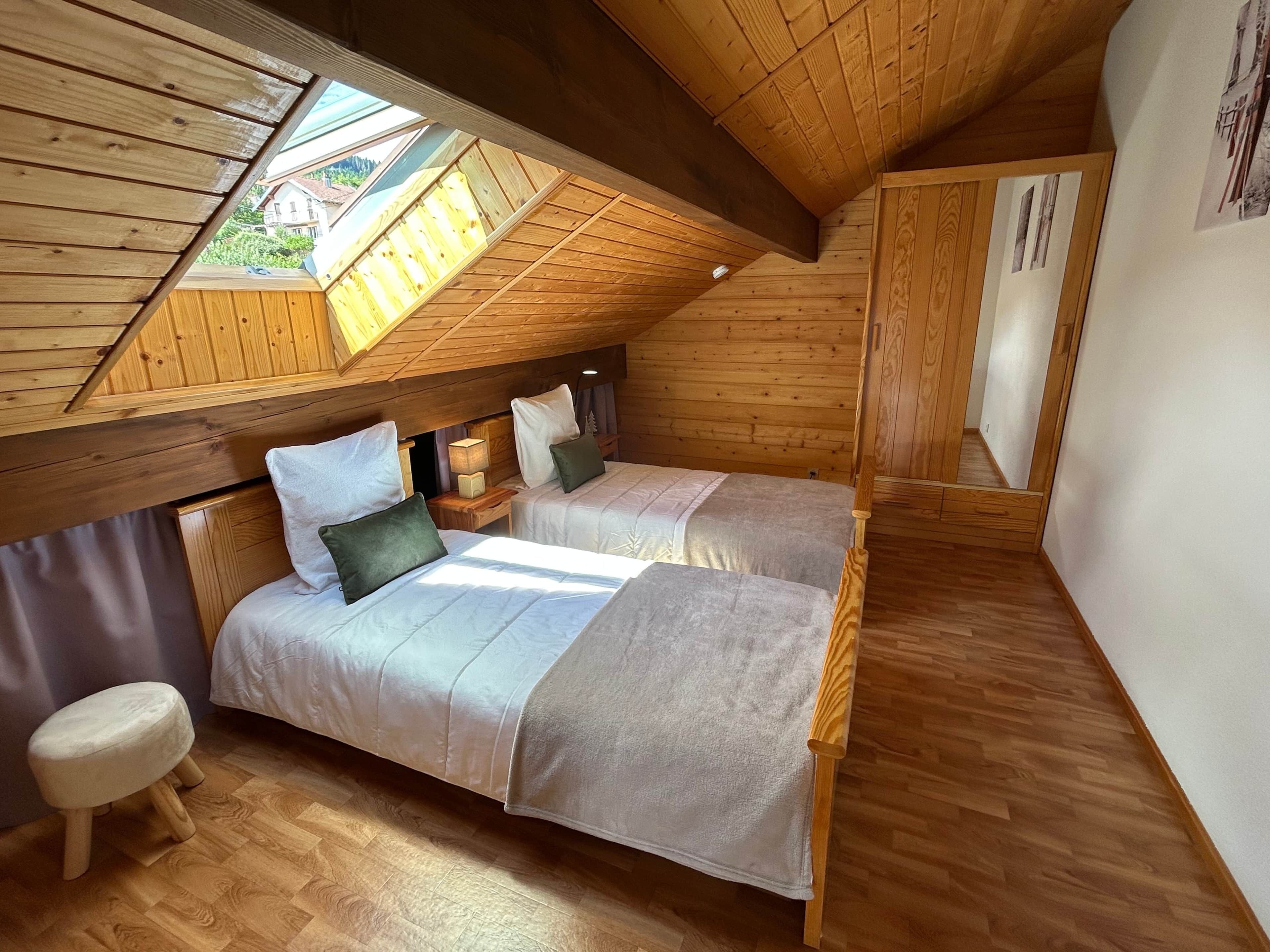A cozy attic bedroom with two beds, wooden beams, a skylight, and a small stool.