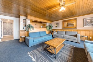 Un salon chaleureux avec un canapé bleu, un canapé gris et une table basse en bois. On y trouve deux tables d’appoint, un téléviseur fixé au mur et un ventilateur de plafond.