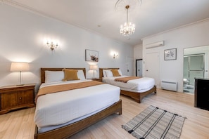 A hotel room with two beds, a chandelier, wall sconces, a wooden floor, and a bathroom visible through an open door.