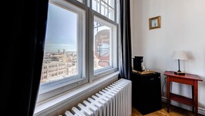 A room with a view of a cityscape through a window, a radiator, a small table with a lamp, and a framed picture on the wall.