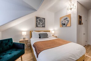 A bedroom with a large bed, a nightstand, a dresser, a green chair, and a chandelier. - Auberge in Old Quebec - 1 bed suite (Québec)