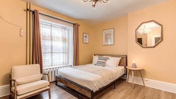Une chambre à coucher comprenant un lit, une chaise, une table de chevet, une fenêtre avec des rideaux, un lustre et des tableaux encadrés accrochés au mur.