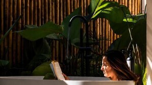 Une personne se détend dans une grande baignoire blanche, un livre à la main, entourée d’une végétation luxuriante et, au fond, de plantes vertes et de bambous.