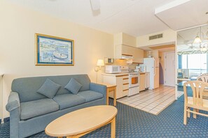 A compact living space with a sofa, a small dining table, a kitchenette with a microwave and stove, and a view of the outside through a door. - Oceanfront Stay by Skywheel! Yachtsman 1002 (Myrtle Beach)