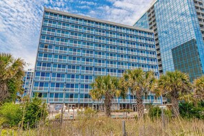 A modern building with a grid of windows, surrounded by palm trees and a fence. - Oceanfront Stay by Skywheel! Yachtsman 1002 (Myrtle Beach)