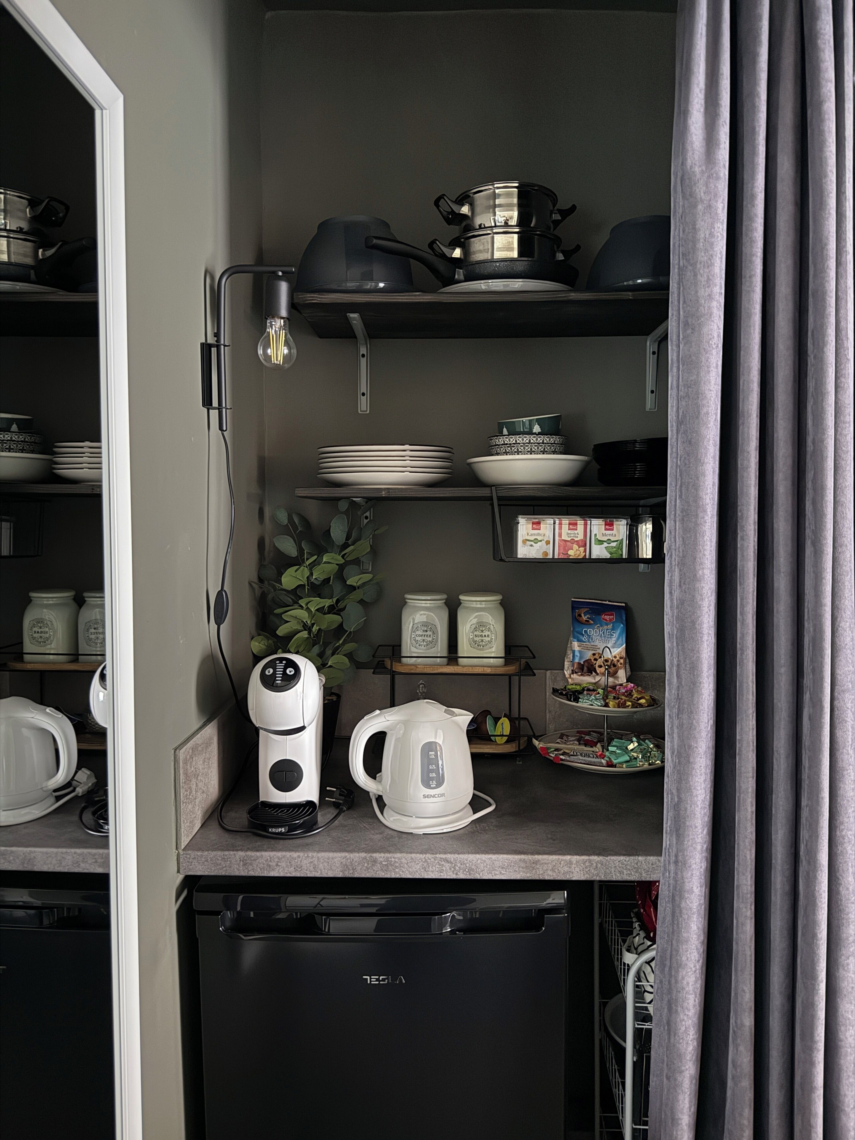 A compact kitchen with a coffee maker, kettle, and shelves holding dishes and jars.