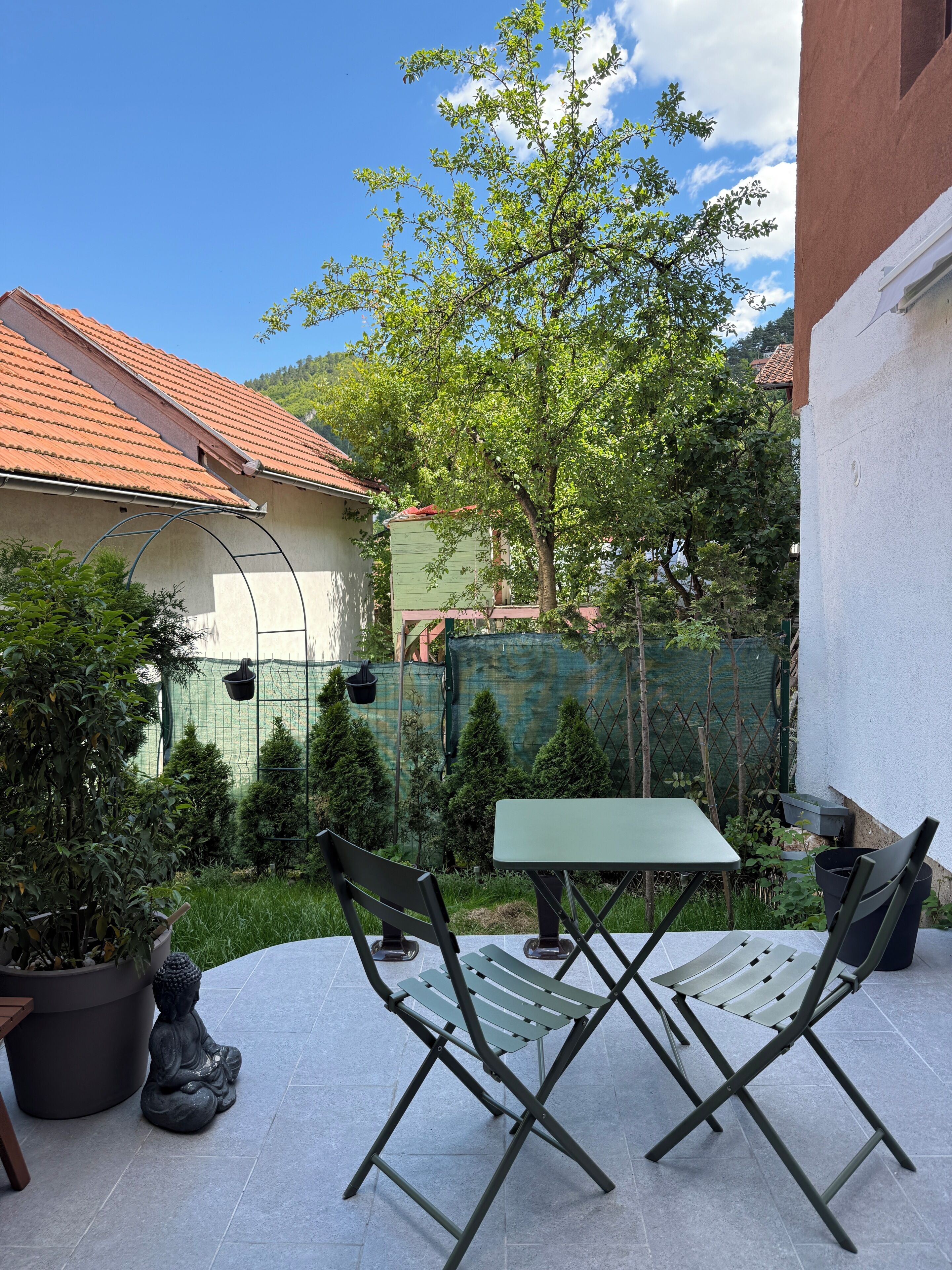 Eine Terrasse im Freien mit Tisch und Stühlen, umgeben von Grünpflanzen und einer Einfriedung.