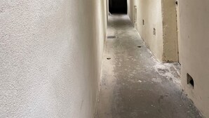 Un couloir étroit aux murs blancs, une porte sombre au fond et une armoire en bois sur la gauche.