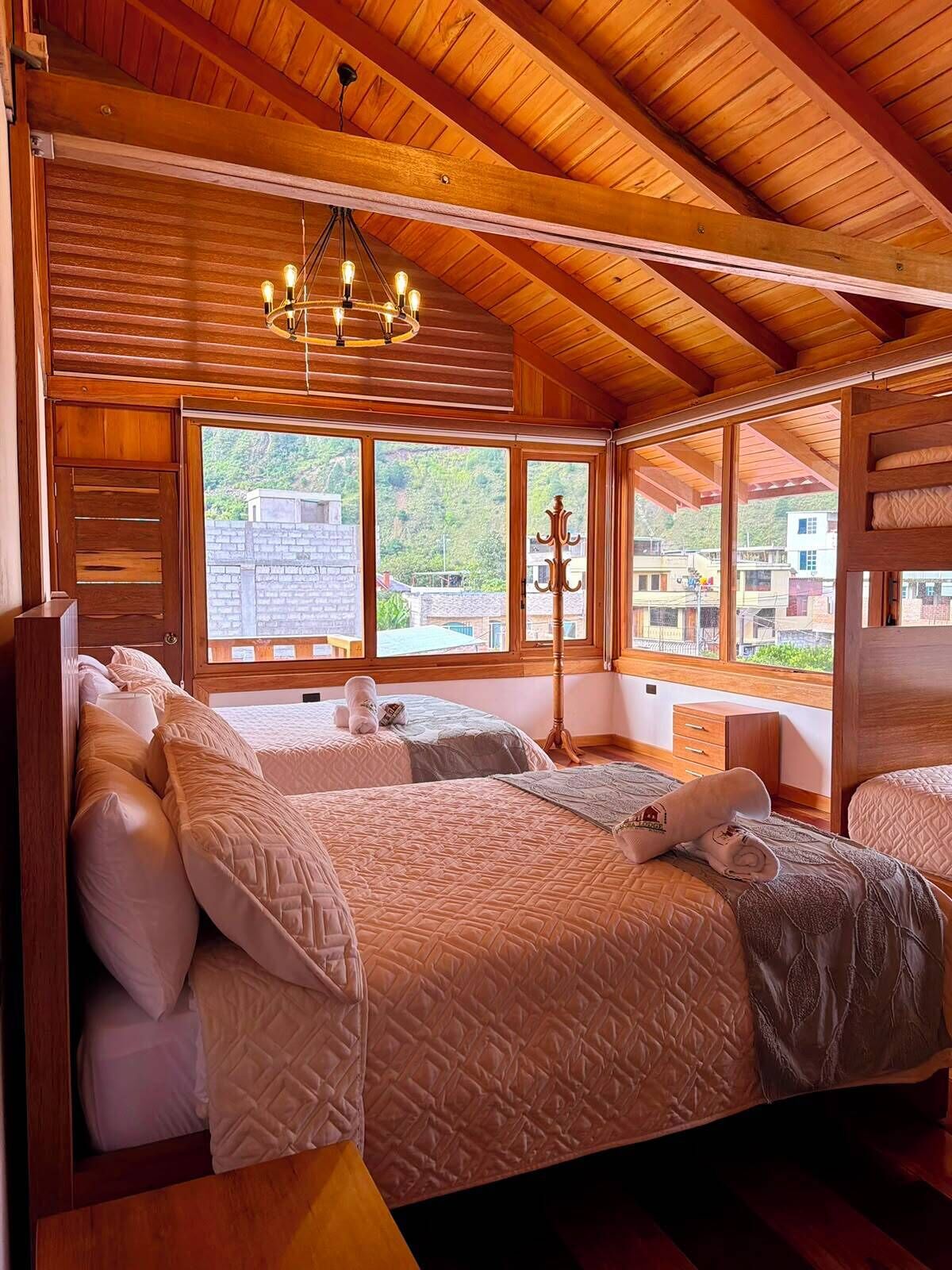 A wooden-paneled room with two beds, a chandelier, and large windows overlooking a hillside.