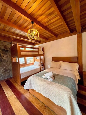 A bedroom with a bunk bed, a single bed, a stone wall, wooden beams, and a hanging light fixture.