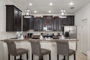 A modern kitchen with dark cabinets, stainless steel appliances, and a bar area with three grey upholstered chairs. - 6Bedroom Luxury Pool Villa near Disney & Free Resort (Four Corners)