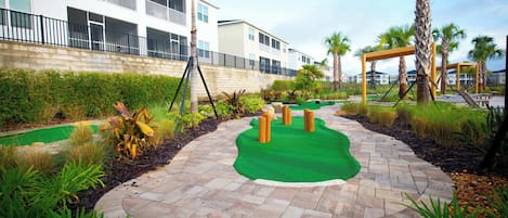 A landscaped pathway with palm trees and a green carpet, leading to a building with balconies.