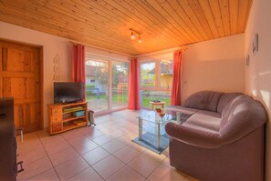 A living room with a television, sofa, glass coffee table, wooden ceiling, sliding glass door, and a view of the outdoors. - Gemutlicher Doppelbungalow mit Terrasse im Ferienpark Mirow (Mirow)