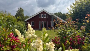 A red building with a gabled roof, surrounded by lush greenery and flowering plants. - Ferienhaus im Skandinavischen Stil mit Garten im Hafendorf Rechlin (Rechlin)