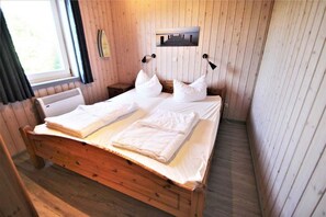 Ein Bett mit Holzrahmen, weiße Bettwäsche und Kissen, ein Nachttisch, ein an der Wand hängendes Bild und ein Fenster mit Vorhängen.