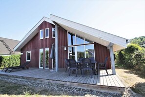 Ein modernes Haus mit Satteldach, großen Fenstern und einer Holzterrasse mit Gartenmöbeln.