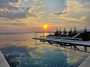 Une piscine à débordement offrant une vue imprenable sur le coucher de soleil sur la mer, des chaises longues et une terrasse en bois.