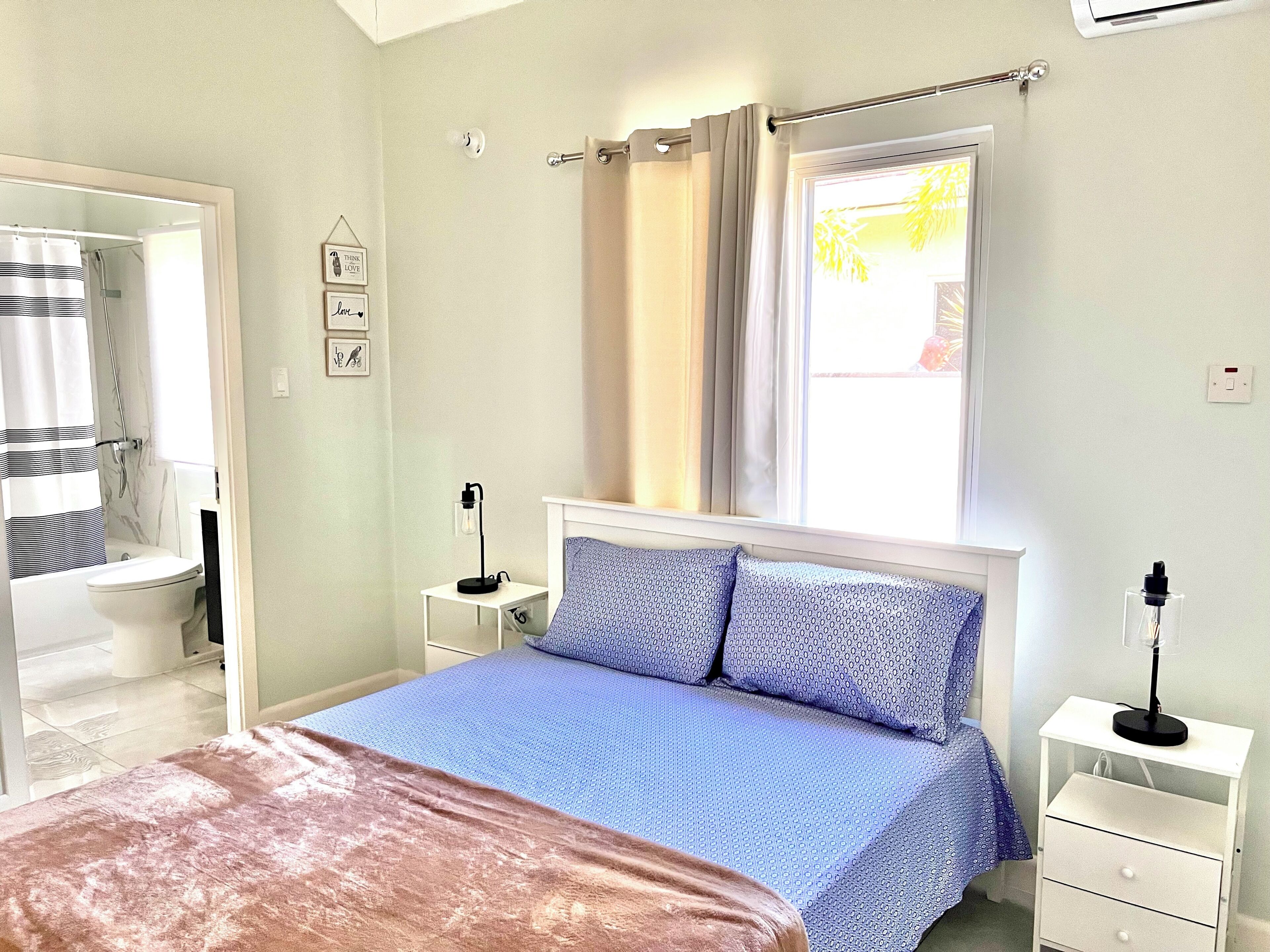 Ein Schlafzimmer mit Bett, Nachttisch, Vorhängen an einem Fenster und einem durchgegangenen Türspalte erkennbaren Badezimmer.