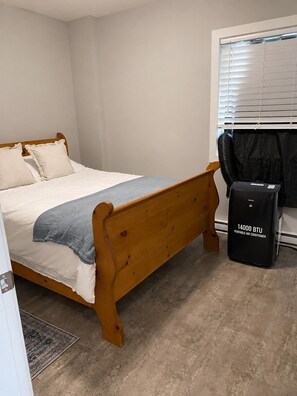 A bedroom with a wooden bed, a window with blinds, and a portable air conditioner.