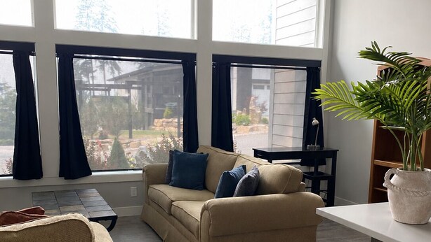 Una sala de estar con un sofá color beige, un sillón oscuro y una mesa de centro. Se ven ventanas amplias con cortinas, una planta en maceta y vistas al exterior.