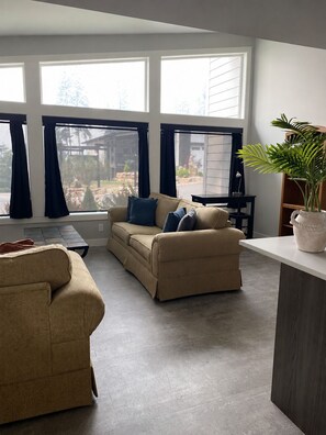 A living room with a beige sofa, a dark-colored armchair, and a coffee table. There are large windows with curtains, a potted plant, and a view of the outdoors.