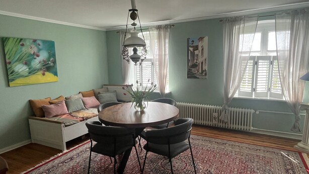 A cozy living room with a round wooden table, black chairs, a white sofa with cushions, a painting on the wall, and a large window with curtains. - Charming vacation rental with garden access, about 4 km from the North Sea (Dagebüll) (Galmsbüll)