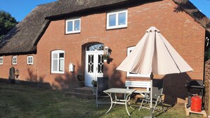A brick house with a thatched roof, a white door, and a small patio with a table and chairs. - Charming vacation rental with garden access, about 4 km from the North Sea (Dagebüll) (Galmsbüll)