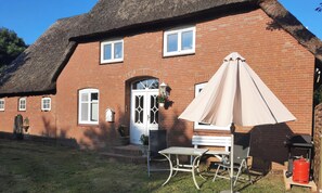 A brick house with a thatched roof, a white door, and a small patio with a table and chairs. - Charming vacation rental with garden access, about 4 km from the North Sea (Dagebüll) (Galmsbüll)