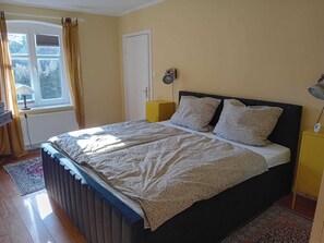 A bedroom with a bed, a window, a door, a bedside table, and a lamp. - Charming vacation rental with garden access, about 4 km from the North Sea (Dagebüll) (Galmsbüll)