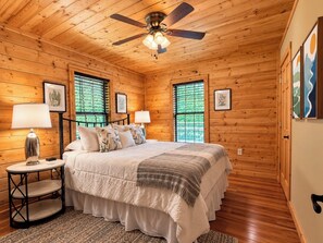 A wooden-paneled bedroom with a bed, two bedside tables with lamps, a ceiling fan, and framed artwork on the walls.