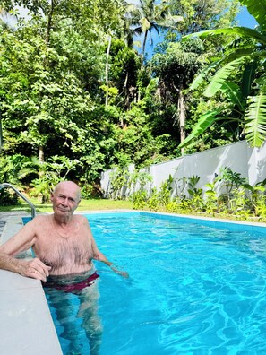 Une piscine extérieure entourée d’une végétation luxuriante et bordée d’un mur blanc.