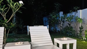 A nighttime outdoor seating area with white lounge chairs and tables, a small tree with white flowers, and a neatly trimmed hedge. - Namo Villa Habaraduwa (Habaraduwa)