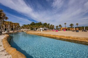 A swimming pool with a curved edge, surrounded by palm trees and a sandy area with lounge chairs. -  One of The Wharf's Newest Vacation Rentals. (Orange Beach)
