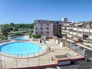 A rooftop pool area with a circular pool, a building with multiple balconies, and a clear sky. - Lagon Azur – Charmant studio avec piscine proche plage (Le Grau-du-Roi)