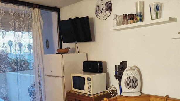 Ett litet kök med mikrovågsugn, brödrost och vattenkokare, ett träbord med en blommig duk och en tv på väggen.