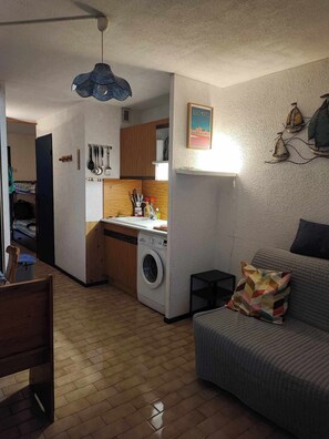 A compact living space with a kitchenette, a washing machine, a dining area, and a sofa with a patterned cushion. - Lagon Azur – Charmant studio avec piscine proche plage (Le Grau-du-Roi)